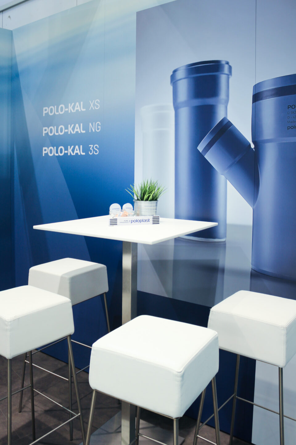 Detailaufnahme des Messestands von POLOPLAST mit Stehtisch, weißen Barhockern und großformatiger Wandgrafik für Rohrsysteme der Serien POLO-KAL XS, NG und 3S.