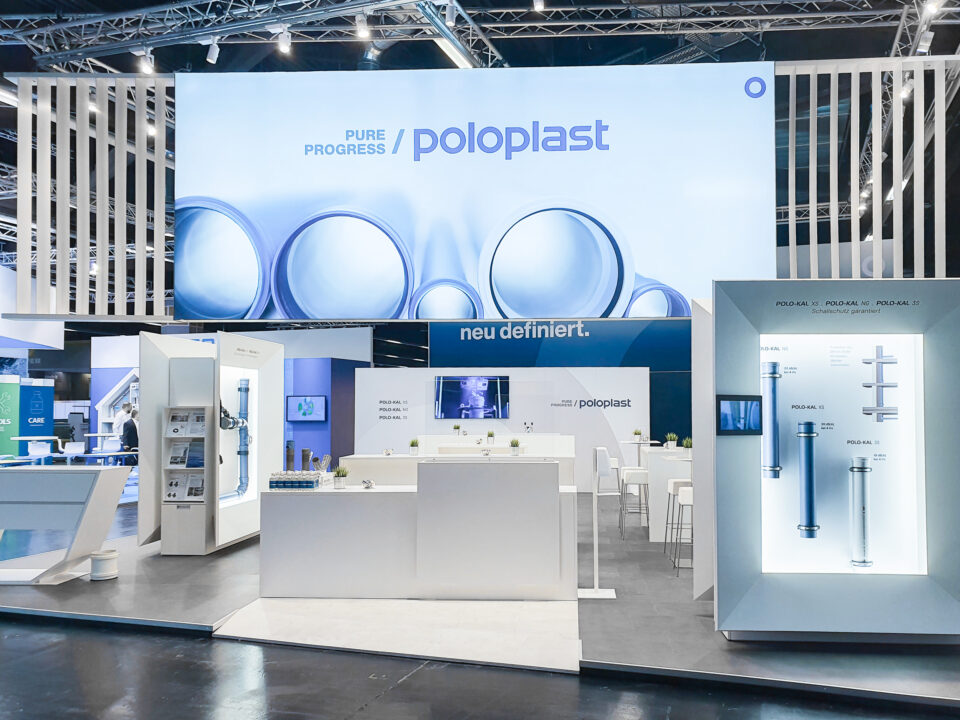 Frontale Perspektive des POLOPLAST-Messestands mit zentralem Thekenbereich, großem LED-Screen und beleuchteten Exponatboxen zur Präsentation der Rohrsystemgenerationen.