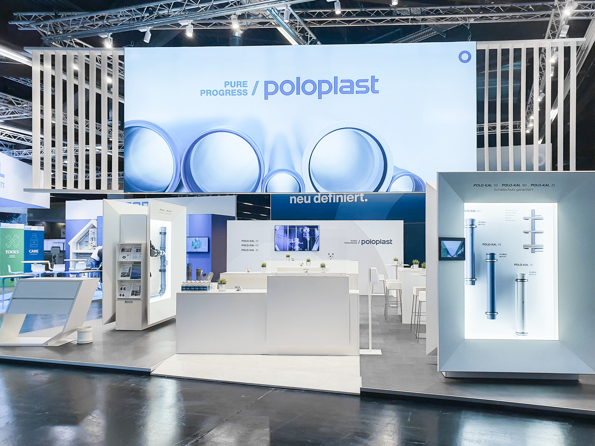 Frontale Perspektive des POLOPLAST-Messestands mit zentralem Thekenbereich, großem LED-Screen und beleuchteten Exponatboxen zur Präsentation der Rohrsystemgenerationen.
