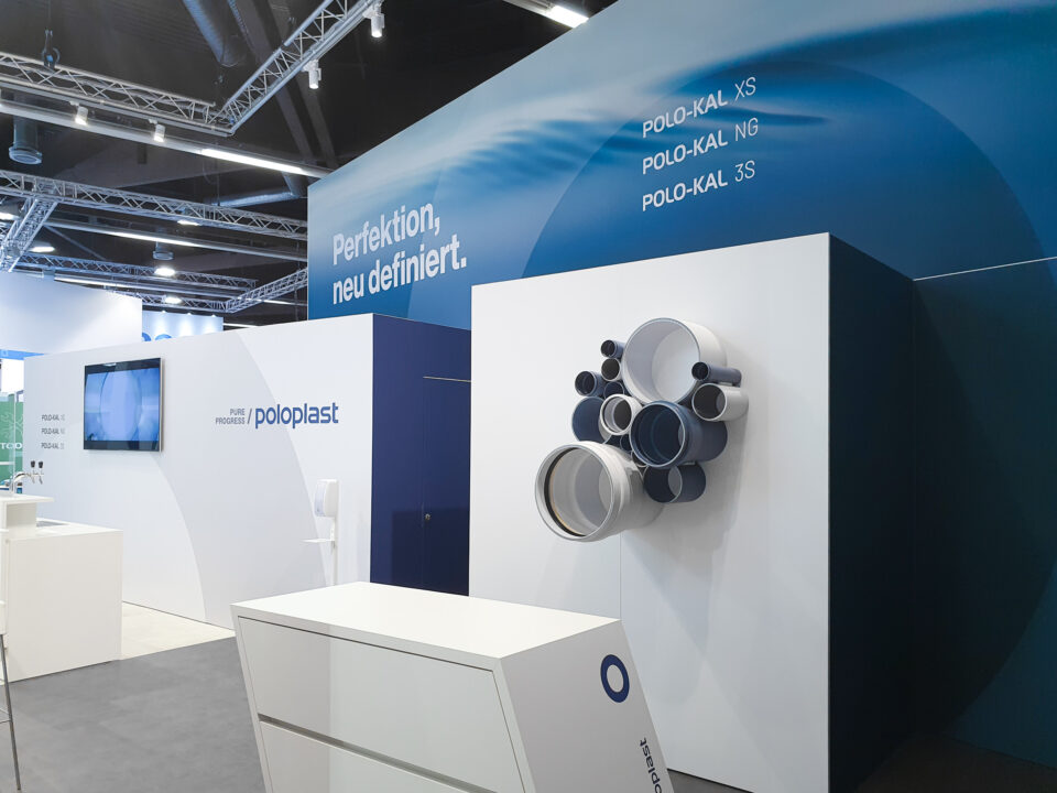 Wandfläche des POLOPLAST-Messestands mit großformatiger Grafik, Claim ‚Perfektion, neu definiert.‘ und skulptural angeordneten Rohrsystemen als visueller Blickfang.